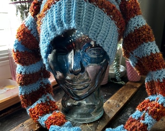 May include: A crocheted hat featuring horizontal stripes of light blue and textured rust-orange yarn. The hat has a ribbed light blue brim and two long, dangling side panels that terminate in light blue, five-pointed star-shaped accents.