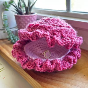 Puede incluir: Un joyero artesanal en forma de concha de crochet rosa intenso. La concha está abierta, revelando un interior lila claro y un anillo con una piedra rosa. La concha está sobre una bandeja de madera cerca de una ventana.