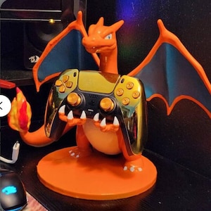 Puede incluir: Una figura naranja de Charizard sosteniendo un mando de videojuegos dorado y negro. La figura tiene alas azules y una llama en la cola. El mando tiene botones y detalles dorados, y descansa sobre una base circular naranja.