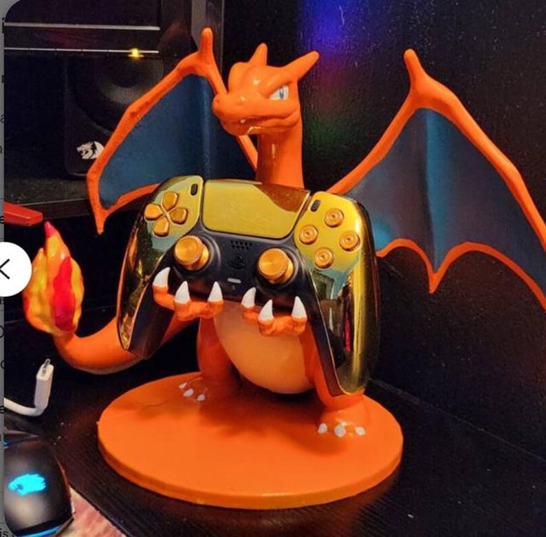 May include: An orange Charizard figure holding a gold and black video game controller. The figure has blue wings and a flame on its tail. The controller has gold buttons and accents, and is resting on an orange circular base.
