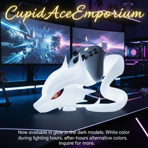 Op de afbeelding: Een witte gaming controller stand in de vorm van een gestileerde drakenkop, met rode ogen, die een zwarte controller vasthoudt. De standaard staat op een zwart bureau met meerdere computermonitors op de achtergrond. De tekst "Cupid Ace Emporium" staat bovenaan.