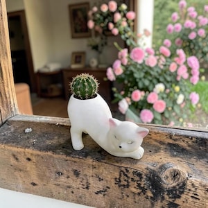 May include: A white ceramic cat-shaped planter with a smooth finish, featuring a small green cactus planted in its back. The cat has closed eyes, a small black nose, and pink-tinted ears, positioned in a stretching pose on a wooden surface.