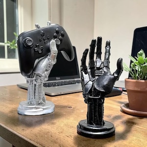 Könnte beinhalten: Zwei roboterartige, handförmige Ständer. Eine silberne Hand hält einen schwarzen Gamecontroller. Die andere ist eine schwarze Hand. Beide stehen auf einer Holzoberfläche. Ein Laptop und eine Topfpflanze sind im Hintergrund.