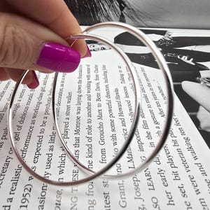 Puede incluir: Un par de pendientes de aro grandes y plateados. Los pendientes son sostenidos por una mano con esmalte de uñas rosa brillante, sobre un fondo de texto impreso y una fotografía en blanco y negro.