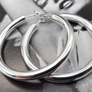 Peut inclure: Deux boucles d'oreilles créoles argentées et brillantes. Les boucles d'oreilles sont épaisses et circulaires, avec une surface lisse et polie. Elles sont présentées sur un fond en noir et blanc.