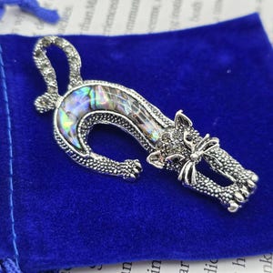 May include: A silver cat brooch with an abalone shell inlay, set on a blue velvet pouch. The brooch has detailed features and a curved tail. The iridescent shell adds a colourful contrast to the silver metal.