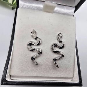 May include: A pair of silver snake earrings, featuring black enamel and clear crystal detailing. The earrings are presented in a black jewellery box with a white interior, highlighting the detailed design and craftsmanship.