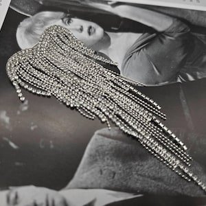 May include: A sparkling silver ear cuff, shaped like a wing, adorned with numerous rhinestones. The design includes a curved top and dangling strands. The cuff is set against a black and white photograph background.