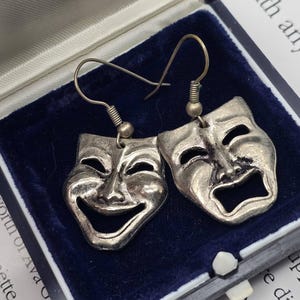 Könnte beinhalten: Silberfarbene Theatermasken-Ohrringe, einer lachend und einer weinend, in einer blauen Samt-Schmuckschatulle präsentiert. Die Ohrringe haben einen Hakenverschluss und detaillierte Gesichtsmerkmale.