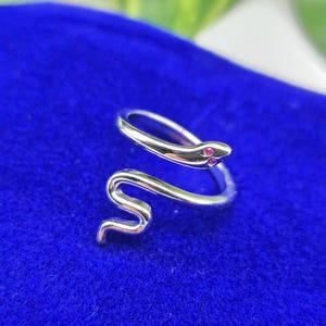 Op de afbeelding: Een zilverkleurige slangenring met een opgerold ontwerp. De kop van de slang heeft twee kleine, rode edelstenen als ogen. De ring staat tegen een levendige blauwe achtergrond, wat een opvallend contrast creëert. De ring is een sieraad.