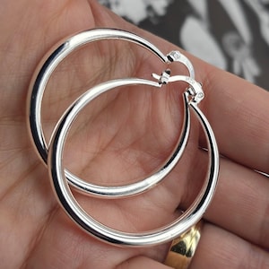 Könnte beinhalten: Ein Paar großer, glänzender silberner Creolen. Die Ohrringe sind rund und haben eine glatte, polierte Oberfläche. Die Reifen werden in einer Hand gehalten, wobei ein goldener Ring an einem der Finger sichtbar ist. Der Hintergrund ist unscharf.