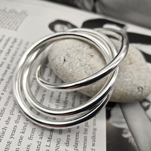 Op de afbeelding: Een set van vier in elkaar grijpende zilveren ringen, waarschijnlijk een armband of een vergelijkbaar accessoire. De ringen zijn glad en reflecterend en liggen op een lichtgrijze steen en een bedrukte pagina.