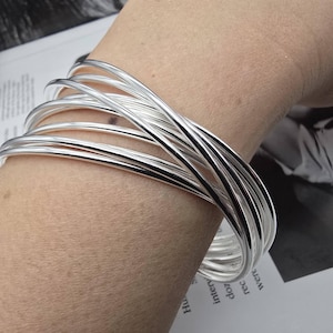 Op de afbeelding: Een stapel glanzende zilveren armbanden om een pols. De armbanden zijn glad en rond en reflecteren licht. De sieraden worden getoond op een lichte huidskleur, met een vage achtergrond van bedrukt papier.