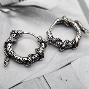 May include: A pair of silver hoop earrings with a snake design. Each earring has a detailed snake wrapped around a textured hoop, resembling a branch. The snakes have textured scales, creating a striking and unique accessory.