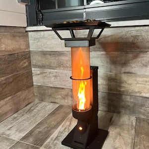 Könnte beinhalten: Ein schwarzer Außenheizer mit einem Glaszylinder, der eine leuchtend orangefarbene Flamme zeigt. Der Heizer hat eine quadratische Basis und eine Oberseite mit einem Grill. Der Heizer steht auf einer gefliesten Oberfläche.