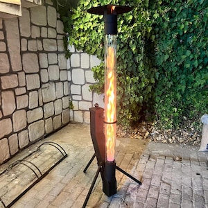 May include: A black outdoor patio heater with a tall, clear glass tube containing a visible flame. The heater has a circular top and is supported by three black legs. The background features a stone wall and green foliage.