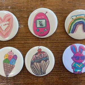 May include: Six circular pinback buttons featuring colorful illustrations: a pink heart with text, a pink digital pet device with the text Feed Me, a rainbow, a multi-colored ice cream cone, a chocolate cupcake with a cherry, and a purple rabbit.