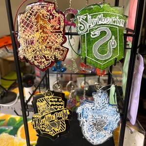 May include: Four resin keychains featuring the crests of Hogwarts houses. The Gryffindor keychain is red and gold, the Slytherin is green and silver, the Hufflepuff is black and gold, and the Ravenclaw is blue and silver. Each keychain has a decorative cord.