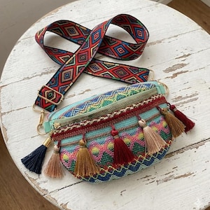 May include: A colorful fanny pack with a patterned strap. The bag features a turquoise base with a zig-zag design and is adorned with tassels in various colors. The strap has a geometric pattern in red, blue, and green.