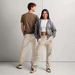 May include: Two people wearing matching beige sweatpants and sneakers. One person wears a brown t-shirt, the other a white crop top and gray zip-up hoodie. The sweatpants have text printed on the left leg.
