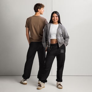 May include: Two people wearing casual outfits. One person is wearing a brown t-shirt and black sweatpants. The other person is wearing a white crop top, gray zip-up hoodie, and black sweatpants. The sweatpants have text on the left leg.