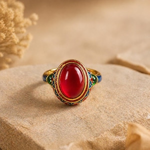 Puede incluir: Un anillo de oro con una piedra preciosa ovalada de color rojo intenso. La banda del anillo tiene detalles de esmalte azul, rojo y verde. La piedra preciosa es el punto focal, engastada en un bisel dorado. El anillo se muestra sobre una superficie texturizada de color marrón claro.