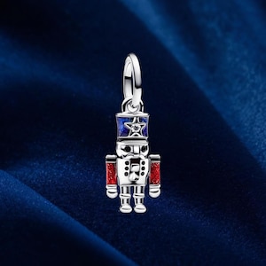 May include: A silver-colored charm in the shape of a toy soldier. The soldier has a blue hat with a star, red arms, and a white uniform. A small, clear gem is set in the hat. The charm is suspended from a silver loop.