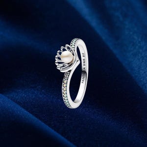 Könnte beinhalten: Ein Ring aus Sterlingsilber mit einer zentralen weißen Perle in einer muschelförmigen Fassung. Die Ringschiene ist mit kleinen, klaren, funkelnden Steinen besetzt. Auf der Innenseite befindet sich die Gravur PANDORA S925 ALE.