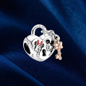 May include: A silver heart-shaped charm with Minnie and Mickey Mouse illustrations. The charm has a key and the words "You & Me" engraved on it. The charm is made of silver and has a rose gold key.