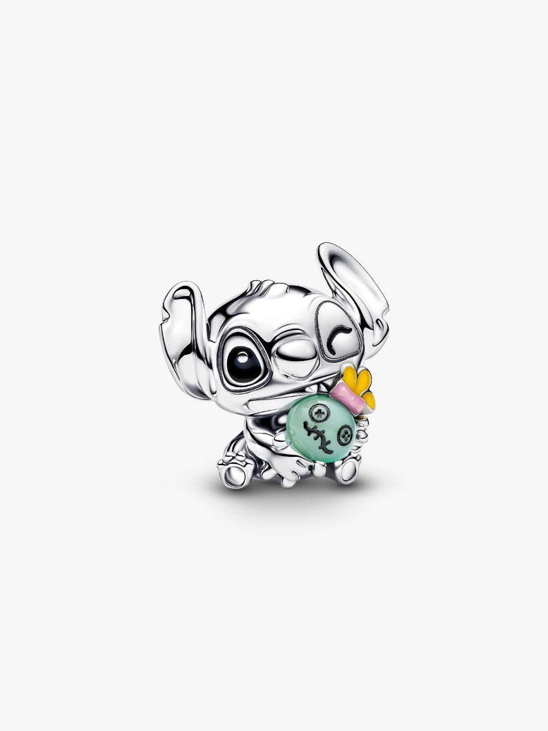 May include: A silver-colored charm featuring the character Stitch from Lilo & Stitch. Stitch is winking and holding a green and yellow charm. The charm is detailed with a cartoon-style design, perfect for Disney fans.