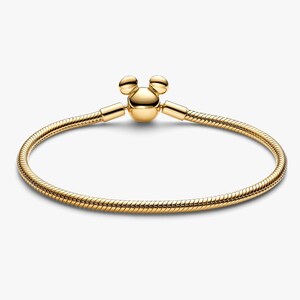 May include: A gold-toned snake chain bracelet featuring a clasp shaped like a stylized mouse head with two rounded ears. The bracelet has a smooth, flexible, and circular design with a polished metallic finish throughout the entire piece.