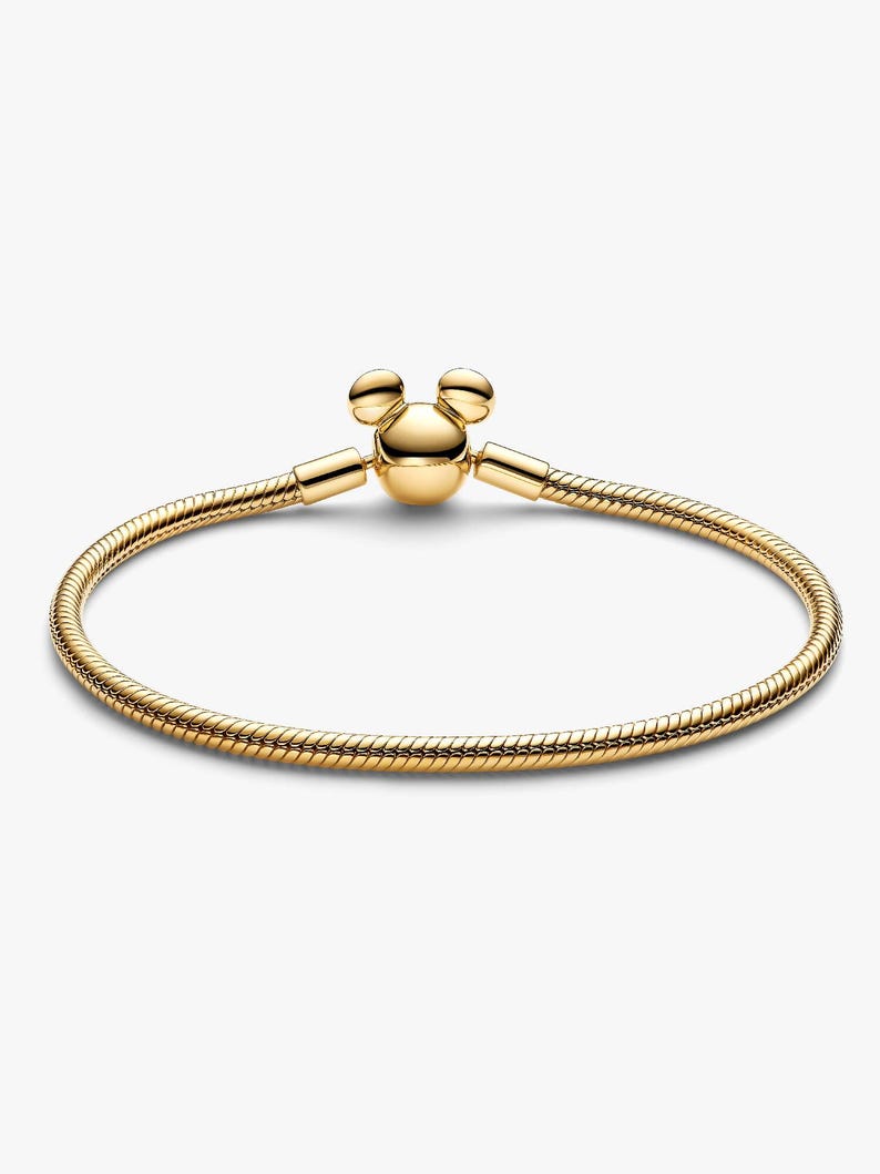 May include: A gold-toned snake chain bracelet featuring a clasp shaped like a stylized mouse head with two rounded ears. The bracelet has a smooth, flexible, and circular design with a polished metallic finish throughout the entire piece.