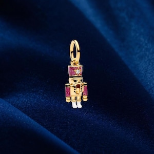 May include: A gold-coloured charm in the shape of a toy soldier nutcracker. The charm has a red hat and arms, white boots, and a small diamond. The background is a dark blue.