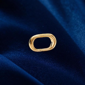 May include: A gold-coloured oval clasp with a hinged opening. The clasp is set against a dark blue background. The clasp is a jewellery component, likely for a necklace or bracelet.