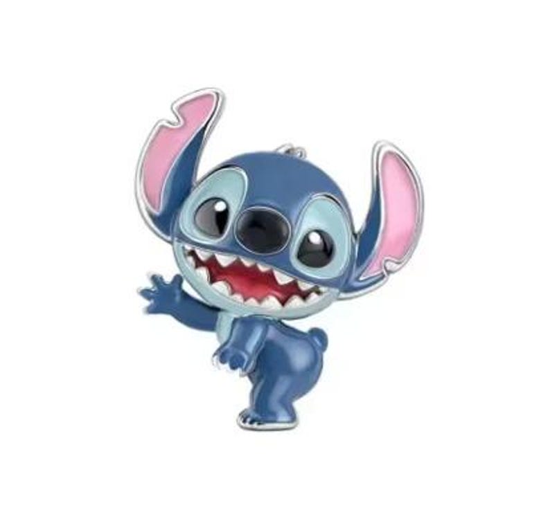 May include: A blue and pink enamel pin featuring the character Stitch from Lilo & Stitch. The pin shows Stitch waving with a wide grin, large black eyes, and pink-lined ears. The pin has a silver outline and is set against a white background.