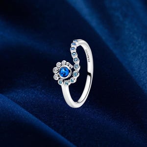 Puede incluir: Un anillo de tono plateado con un diseño en espiral que termina en un grupo circular de piedras. El anillo tiene una piedra central azul oscuro rodeada de piedras más pequeñas de color azul claro y transparentes, con la marca PANDORA grabada.