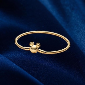 May include: A gold-toned snake chain bracelet featuring a central clasp shaped like a stylized mouse head with two rounded ears. The bracelet has a smooth, polished finish and a flexible, circular design that maintains its shape.