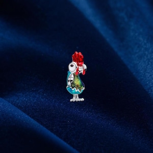 Puede incluir: Un charm de gallo de esmalte colorido con cresta roja, ojos blancos y cuerpo azul y verde. El charm es de metal color plata y se apoya sobre dos pequeños pies. El gallo es un diseño de estilo caricatura.