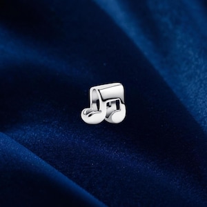 Peut inclure: Un charm argenté brillant en forme de croche. La surface du charm est lisse et réfléchissante, sur un fond bleu foncé. Ce bijou symbolise la musique et la créativité.
