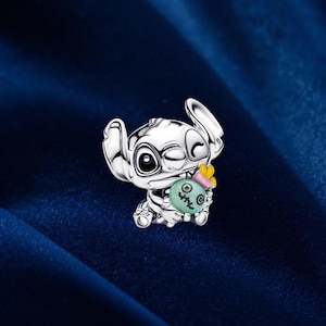 May include: A silver-colored charm featuring the character Stitch from Lilo & Stitch. Stitch is winking and holding a green object with a pink bow and yellow flower. The charm is set against a dark blue background.