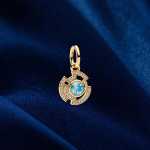 Puede incluir: Un colgante dorado con una piedra central azul claro, rodeada de pequeñas piedras transparentes. El colgante tiene un diseño circular con un patrón decorativo y un lazo para una cadena.