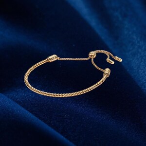May include: A gold bracelet with a textured, beaded design. The bracelet has an adjustable sliding clasp. The jewellery is displayed on a dark blue velvet surface.