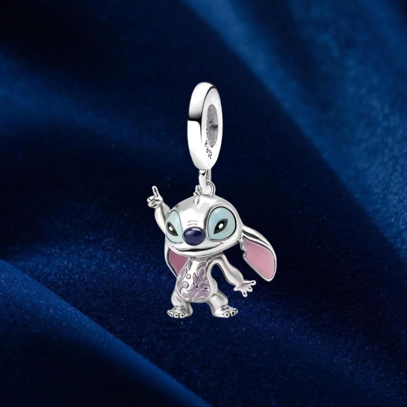 May include: A silver charm featuring the character Stitch from Lilo & Stitch. The charm has a detailed design with blue eyes, pink ears, and a purple floral pattern on its body. The charm is suspended from a silver loop.