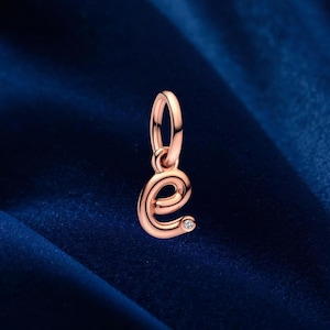 Op de afbeelding: Roségouden bedel in de vorm van de letter "e". De bedel heeft een kleine, heldere edelsteen ingebed in de onderste curve. De bedel is opgehangen aan een kleine lus.