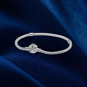 Puede incluir: Una pulsera de cadena de serpiente plateada con un cierre redondo y texturizado que presenta grabados intrincados inspirados en estrellas y el cielo. La pulsera tiene un diseño de metal tejido flexible y un cierre integrado.