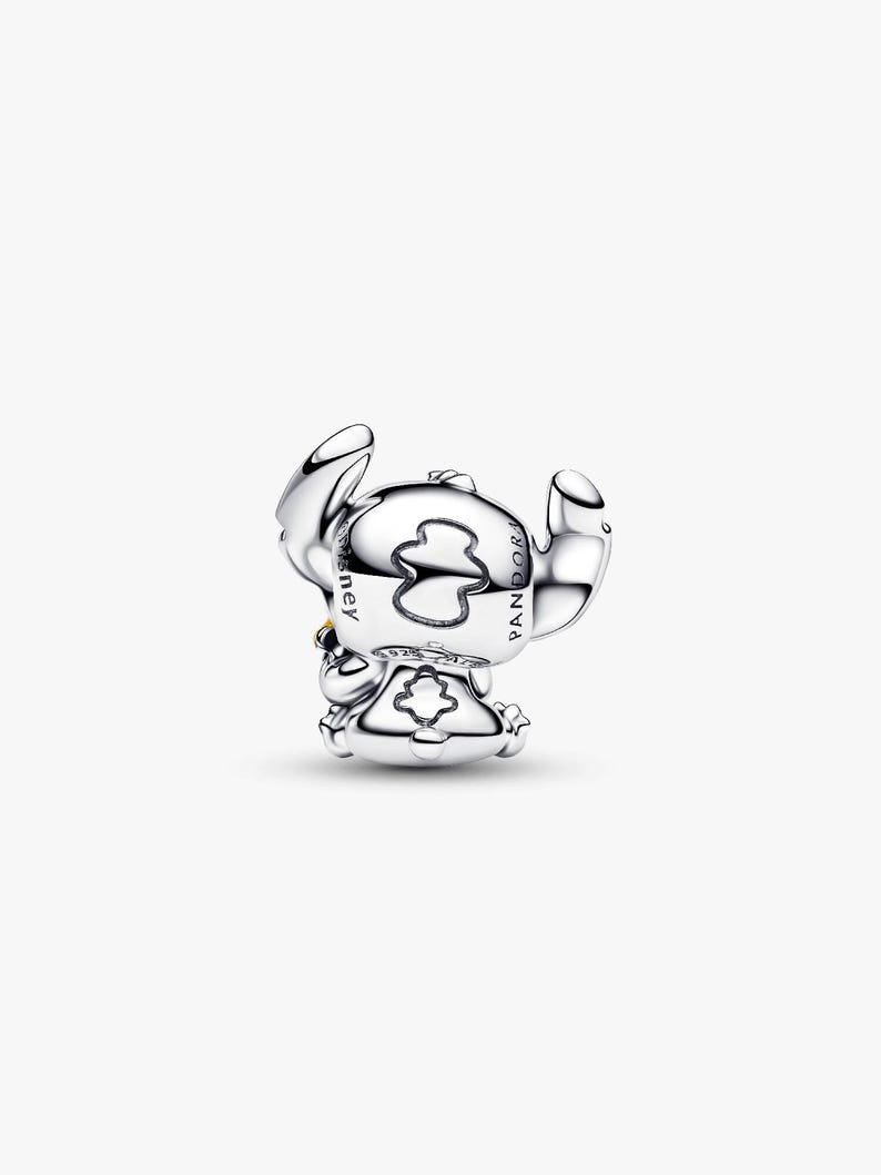May include: A silver charm depicting the character Stitch from the Disney movie Lilo & Stitch. The charm is in a sitting pose with large ears and a small yellow detail. The charm is marked with the Disney and Pandora logos.