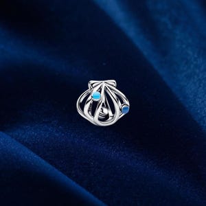 May include: A silver seashell charm with two blue accents. The charm is made of a shiny metal and has an open design. The background is a dark blue fabric.