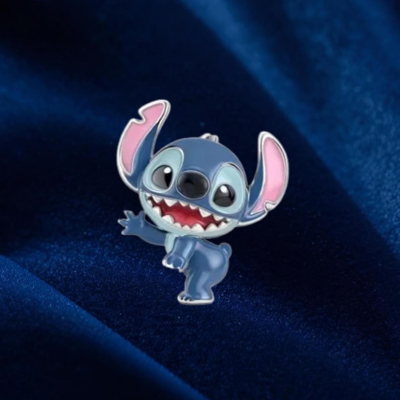 May include: A blue enamel pin featuring the character Stitch from Lilo & Stitch. The pin has a silver outline, pink inner ears, and a wide, toothy grin. The character is in a waving pose.