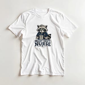 Peut inclure: Un t-shirt blanc à manches courtes avec un motif de raton laveur fatigué portant une tunique bleue et un stéthoscope. Le raton laveur tient une tasse avec le texte CAFFEINE BECAUSE SLEEP IS OPTIONAL. En dessous, le texte NIGHT SHIFT NURSE.