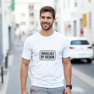 Peut inclure: T-shirt blanc à col rond avec le texte "UNAVAILABLE BY DESIGN" dans un design rectangulaire noir et blanc. Le t-shirt est porté.